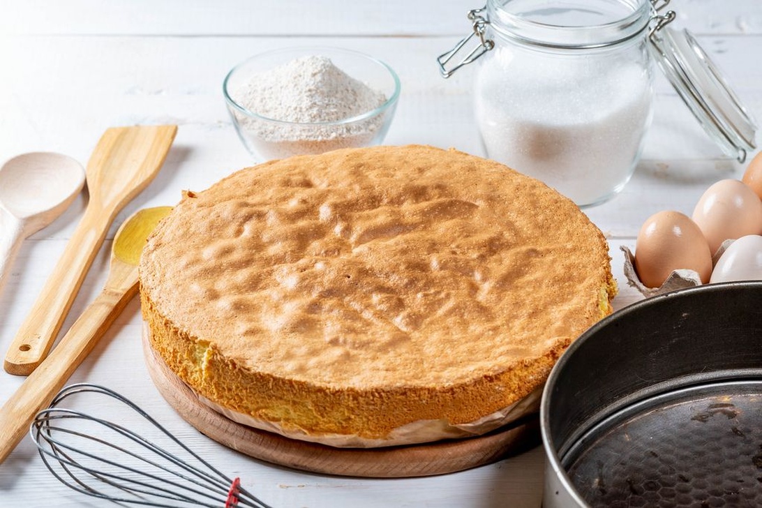 En nygräddad sockerkaka på en träskärbräda omgiven av bakingredienser som ägg, mjöl, socker i en burk och träredskap, vilket illustrerar hemmabakningens charm och grundläggande komponenter.